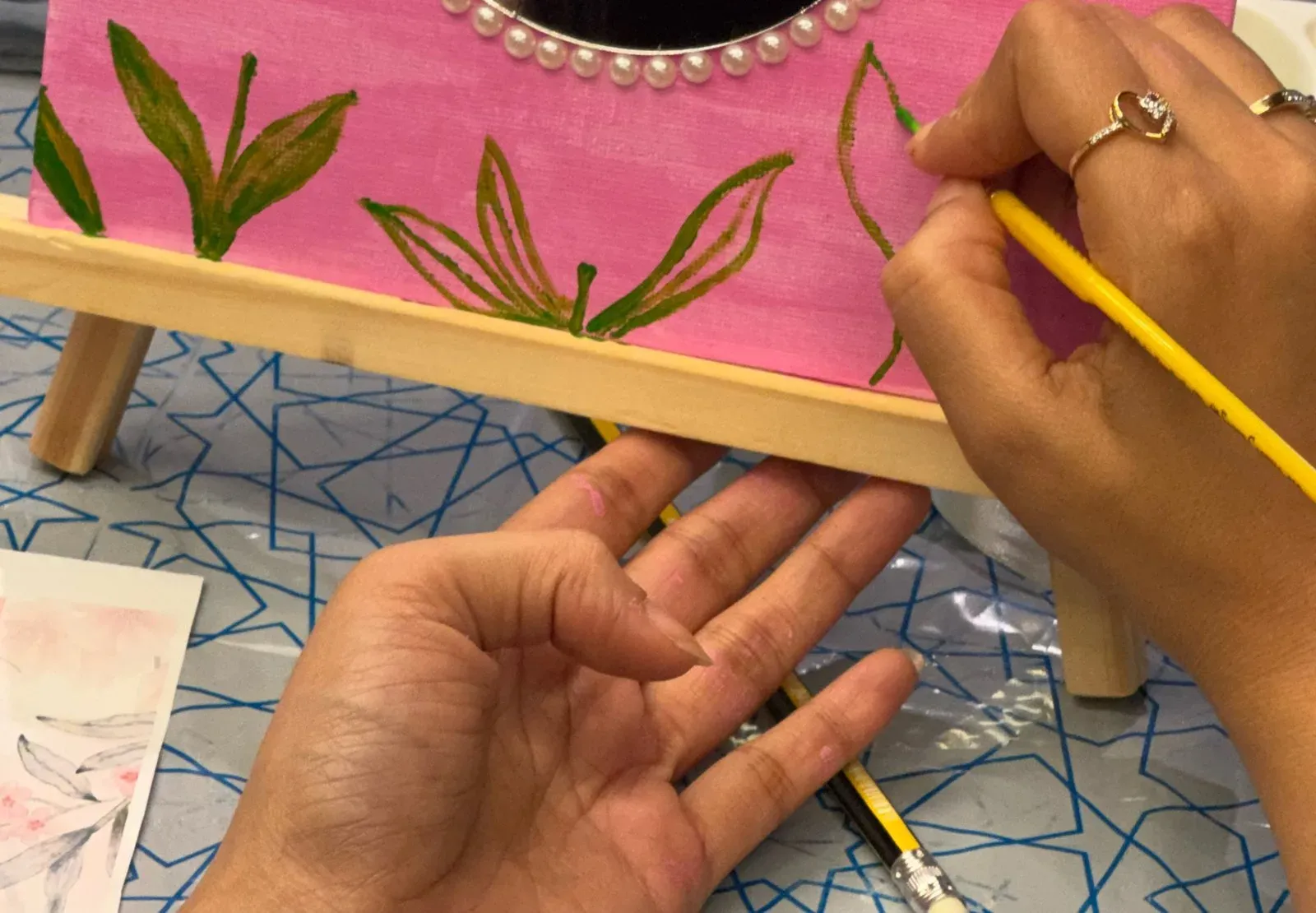 Hands painting green tulip leaves on a pink mirror frame on a small easel, with pearls along the edge of the mirror