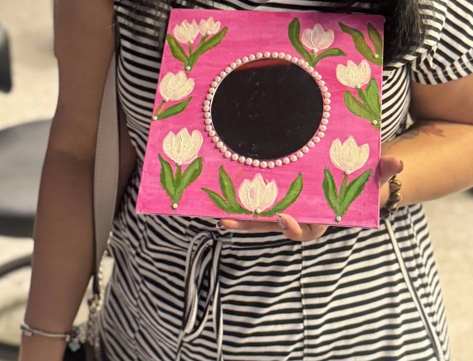 A finished pink mirror frame held up at Moshi, painted with white and pink tulips and green leaves, with a ring of pearls around the round mirror