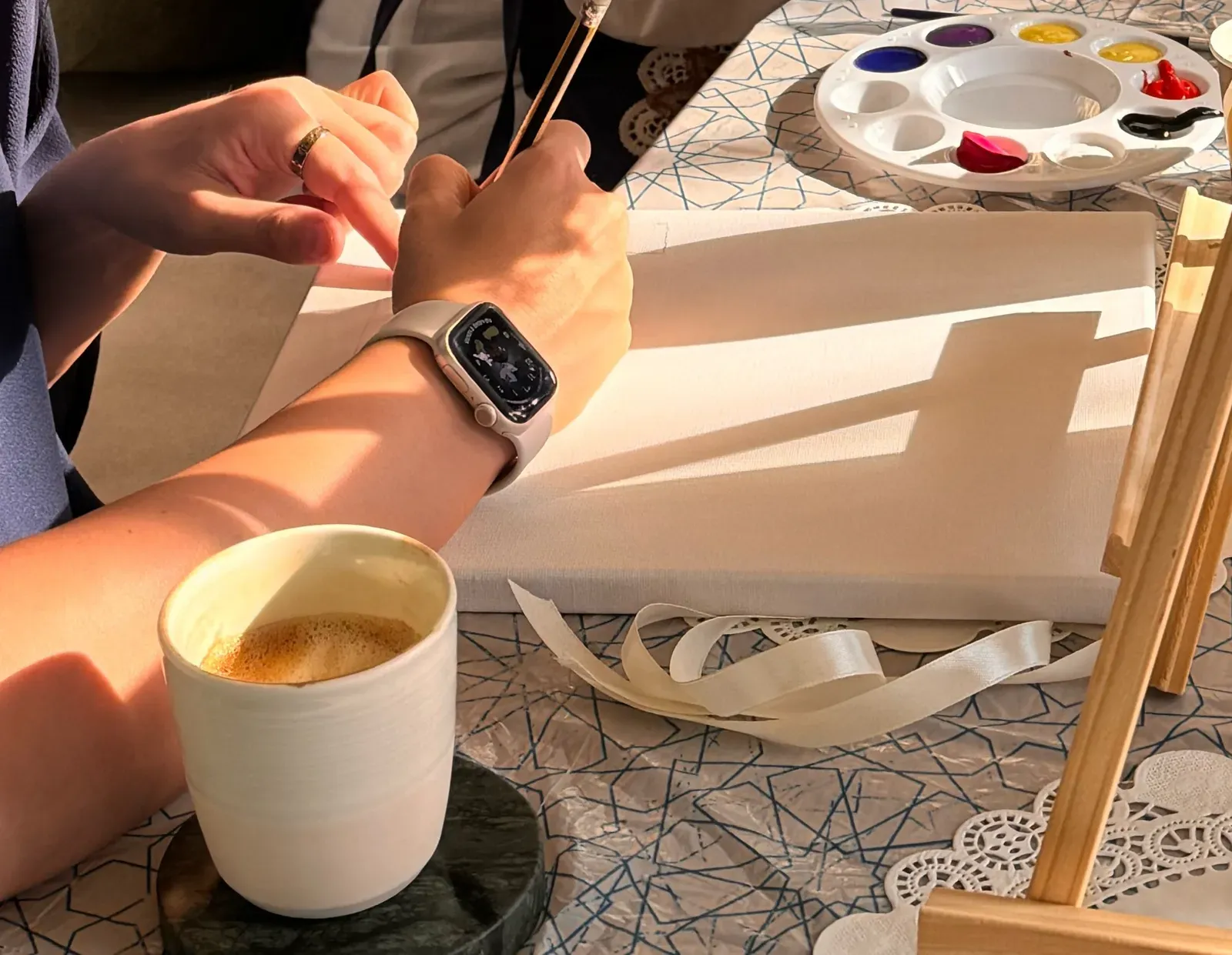 Hands of a guest holding a brush over a blank canvas, with coffee and a palette nearby