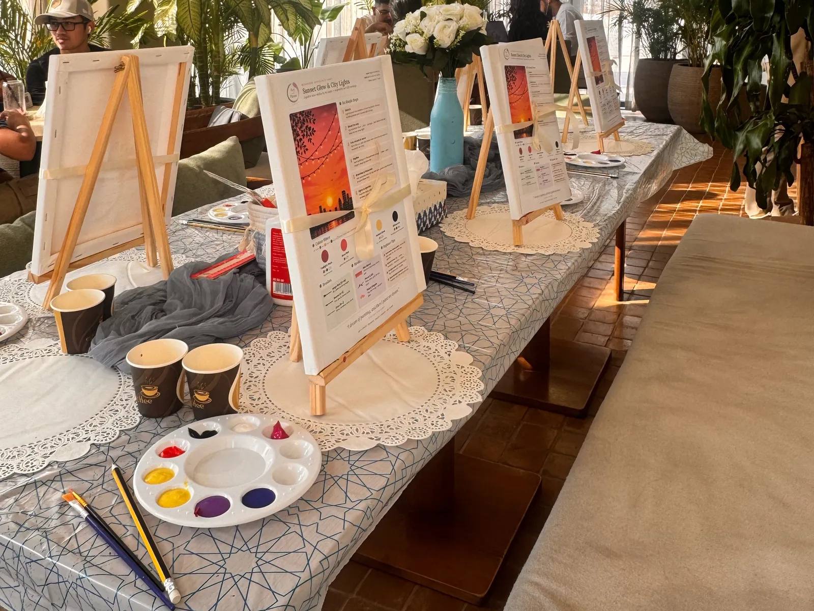 Long table at Ritual Cafe set for the Sunset Glow and City Lights workshop, with blank canvases on wooden easels, printed sunset reference cards, paint palettes and white roses